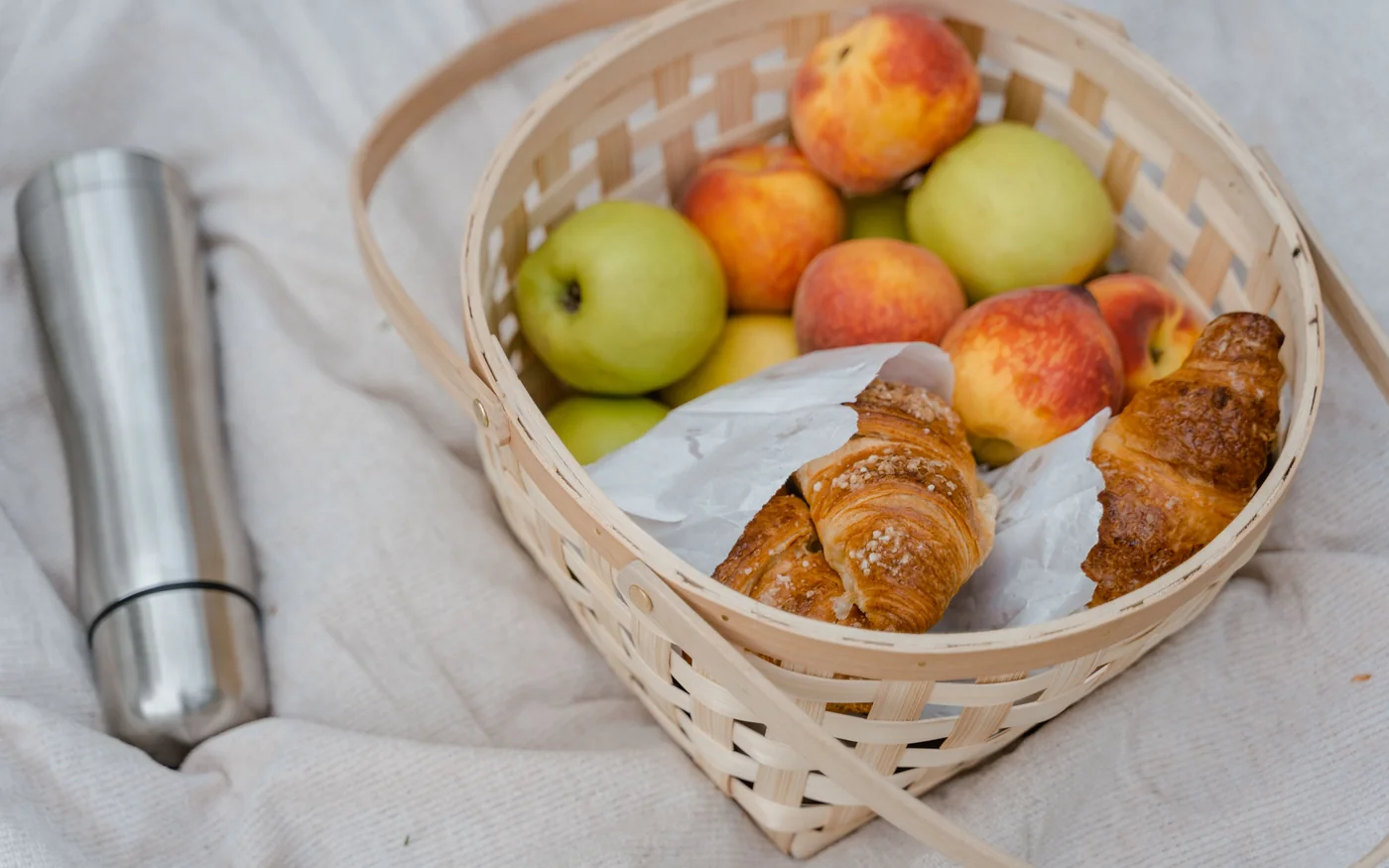 Korb mit ganzen Äpfeln, Pfirsichen und Croissants neben einem Thermosbecher auf einer Picknickdecke