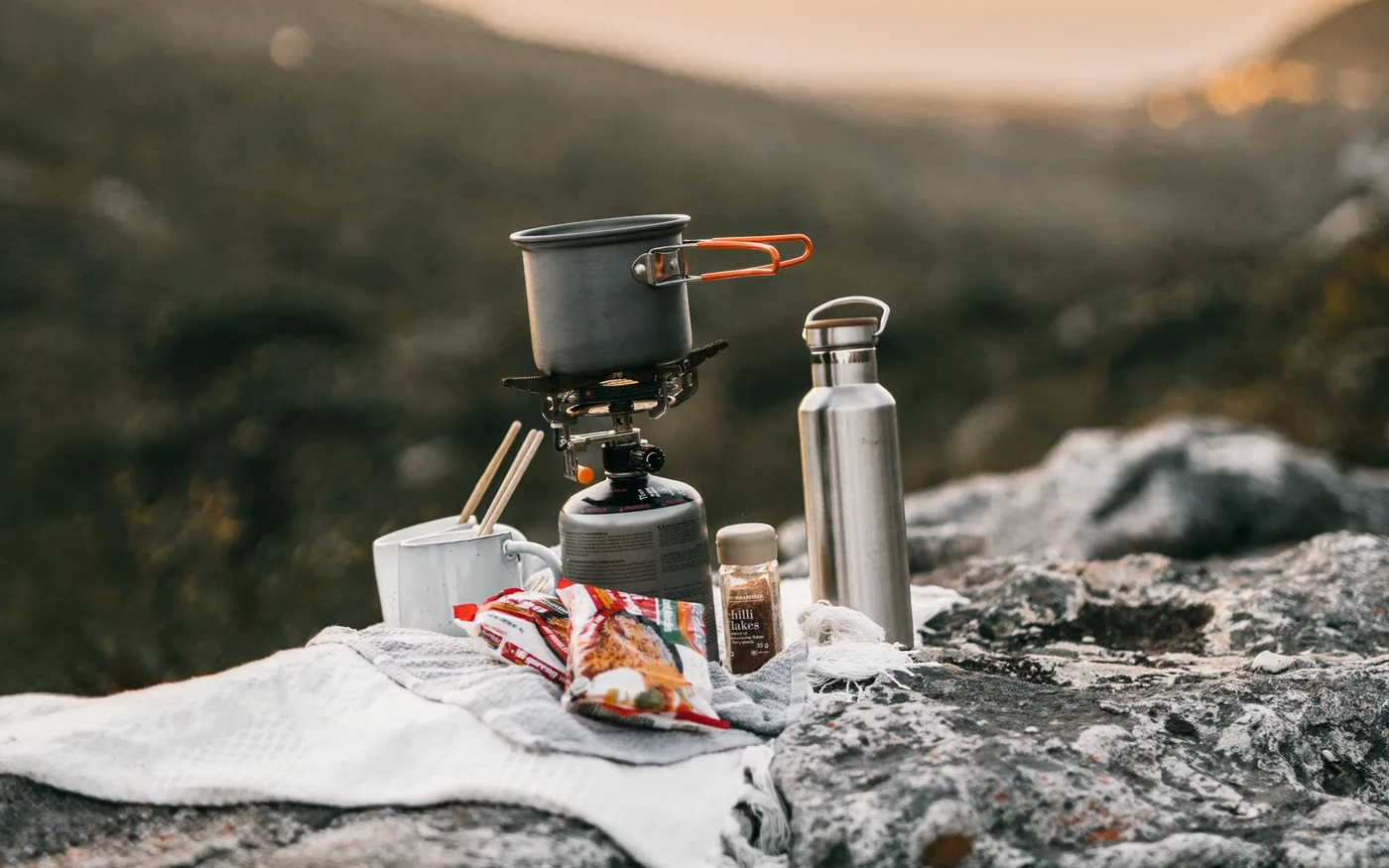 Campingkocher mit Topf, Trinkflasche und Becher auf Felsen im Abendlicht