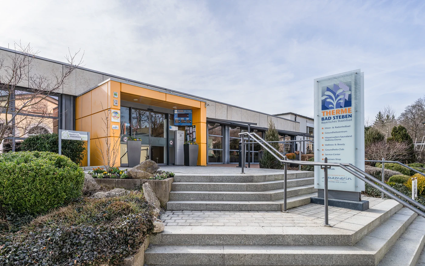 Eingang der Therme Bad Steben mit orangefarbener Fassade und Hinweisschild im Kurpark
